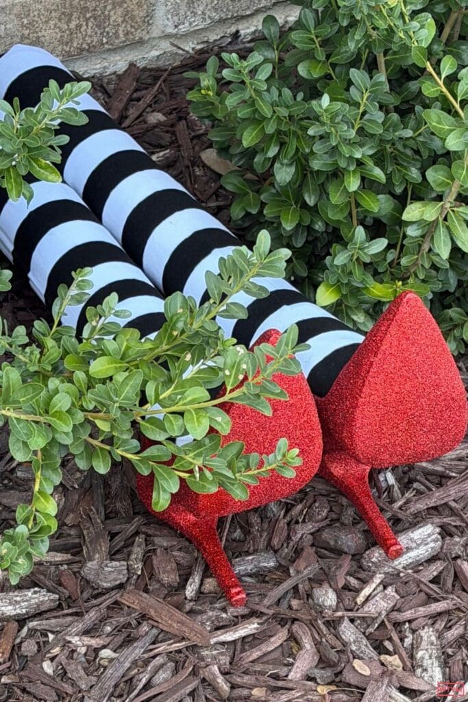 DIY witch legs with striped tights and red glitter ruby slippers lying in mulch against a brick house, like the Wizard of Oz witch under the house